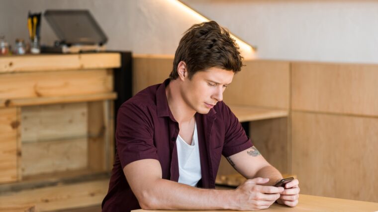 handsome young man using smartphone while sitting at table in cafe